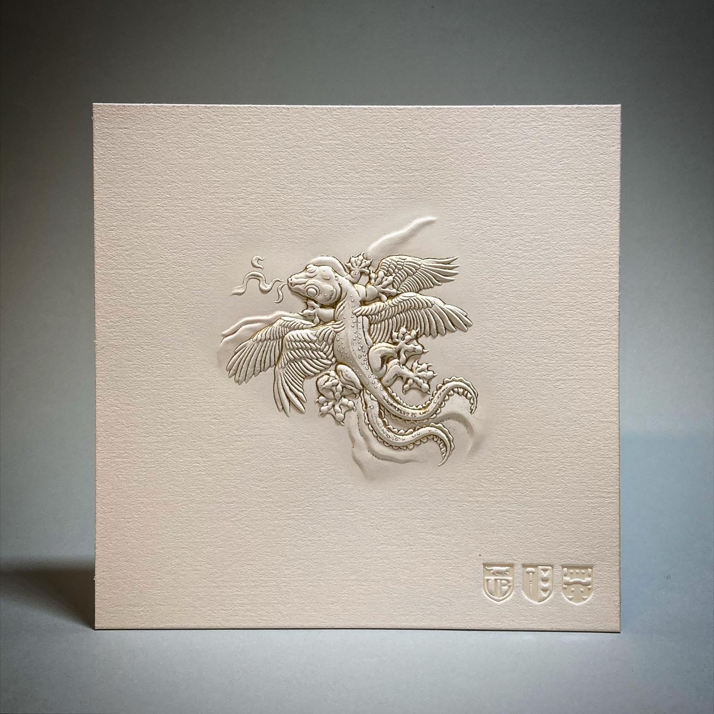 Carte carrée en papier texturé portant un gaufrage en relief d'un lézard ailé aux plumes détaillées et à la queue enroulée, avec trois petits blasons gaufrés dans le coin inférieur droit